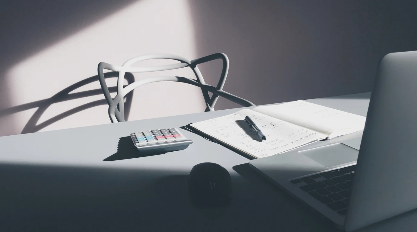 A pale grey desk in hard window light: a calculator, an open notebook with a pen and the edge of a laptop.