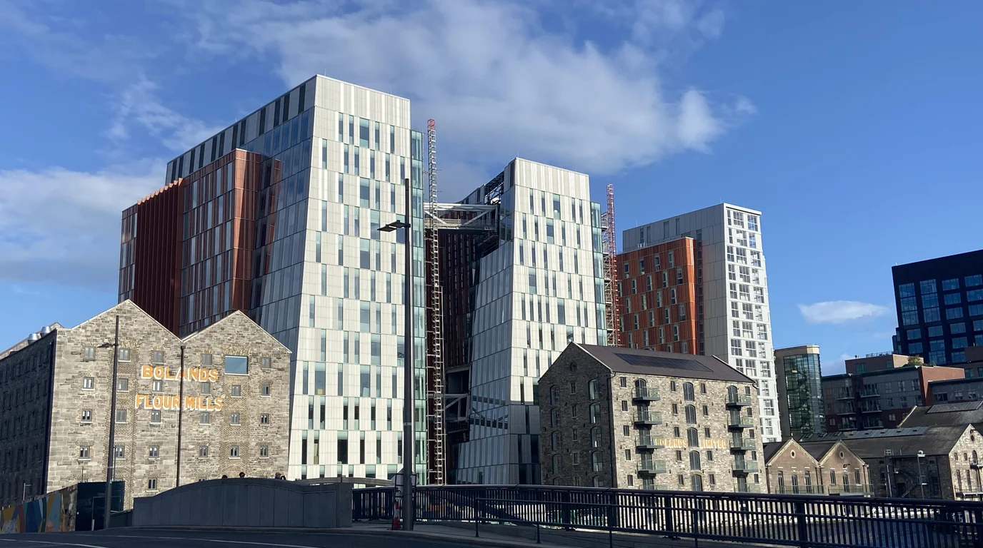 Old stone flour-mill warehouses in front of new glass office towers on Dublin's docklands under a blue sky.