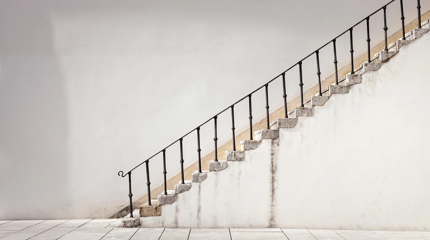 A simple staircase with a black iron handrail climbing along a pale plastered wall.