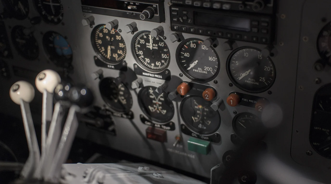 An aircraft instrument panel of round analogue gauges, with white-knobbed throttle levers in the foreground.