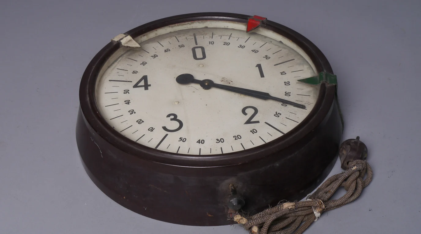 An old electric chronometer with a large white dial and red and green markers, on a grey background.