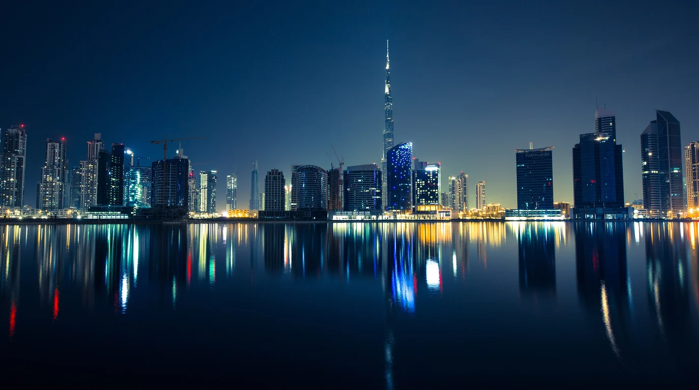 Dubai's skyline at night, the Burj Khalifa rising above the towers, all reflected in still water.