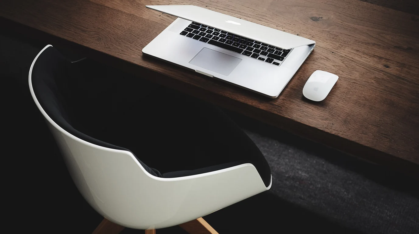 A closed laptop and a mouse on a dark wooden desk, an empty white chair pulled up to it.