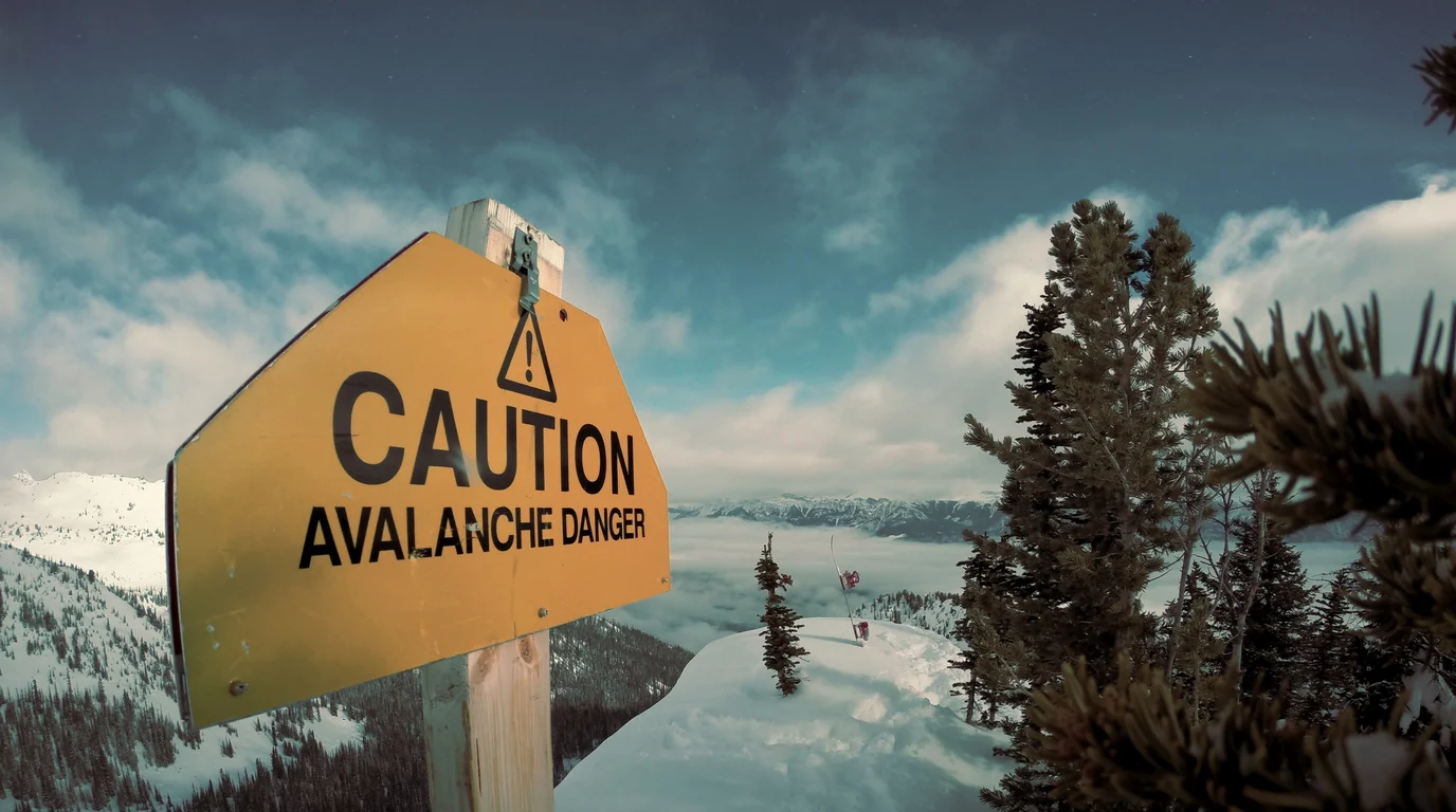 A yellow avalanche-danger warning sign on a snowy mountain slope, with pine trees and low cloud behind it.