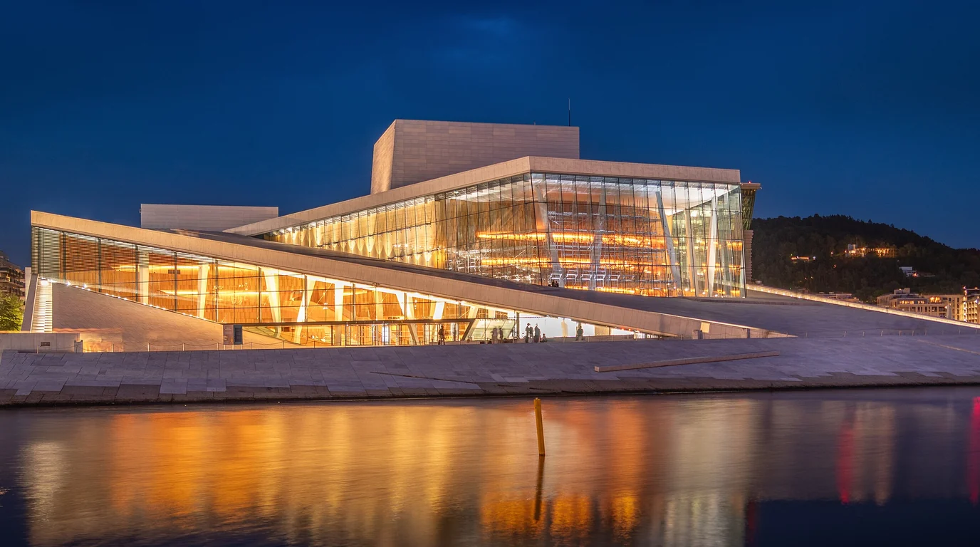 The Oslo Opera House lit up at dusk, its white roof and glass front reflected in the fjord.