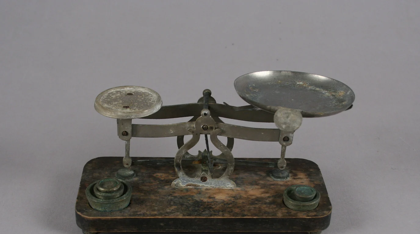 A small antique apothecary balance with two pans and tiny brass weights on a dark wooden base.