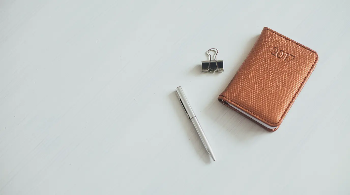 A small tan leather pocket diary next to a silver pen and a black binder clip on a white desk.