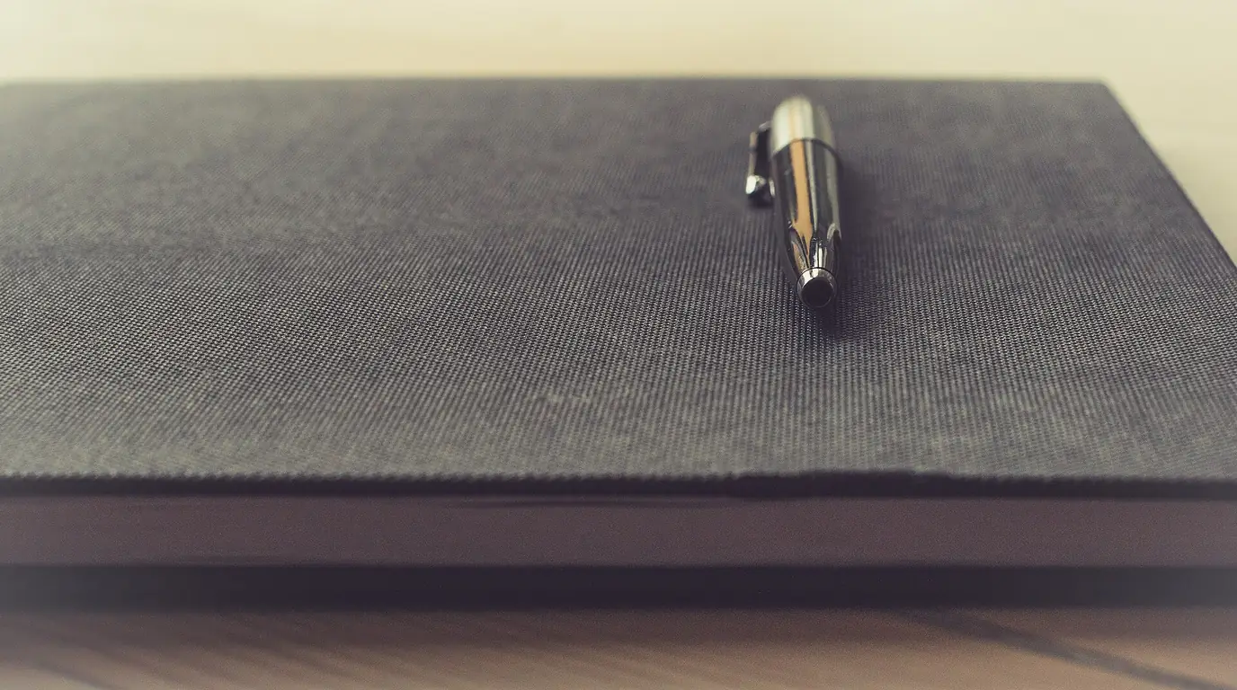 A dark fabric-covered notebook on a desk with a silver ballpoint pen lying across it.