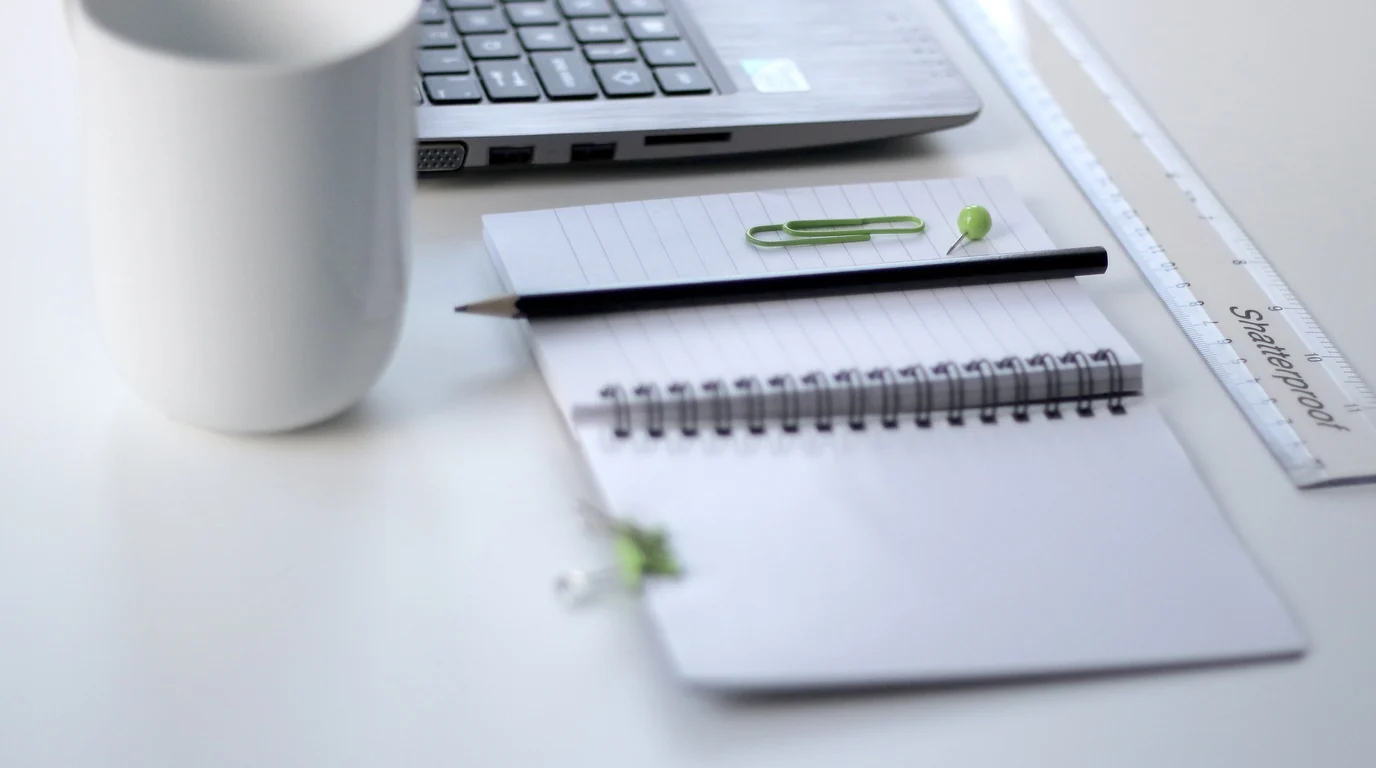 A white desk with a mug, a notebook, a pencil and a clear ruler beside a laptop keyboard.
