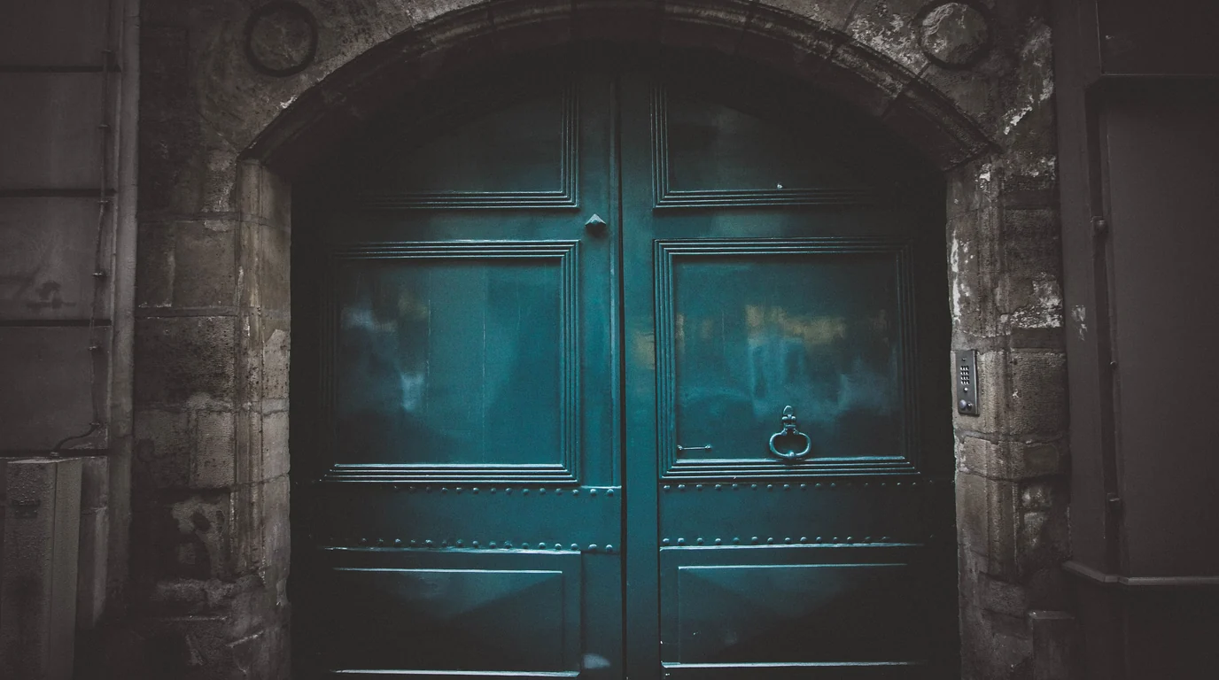A tall teal double door with an iron ring knocker, set in a weathered stone archway in Paris.