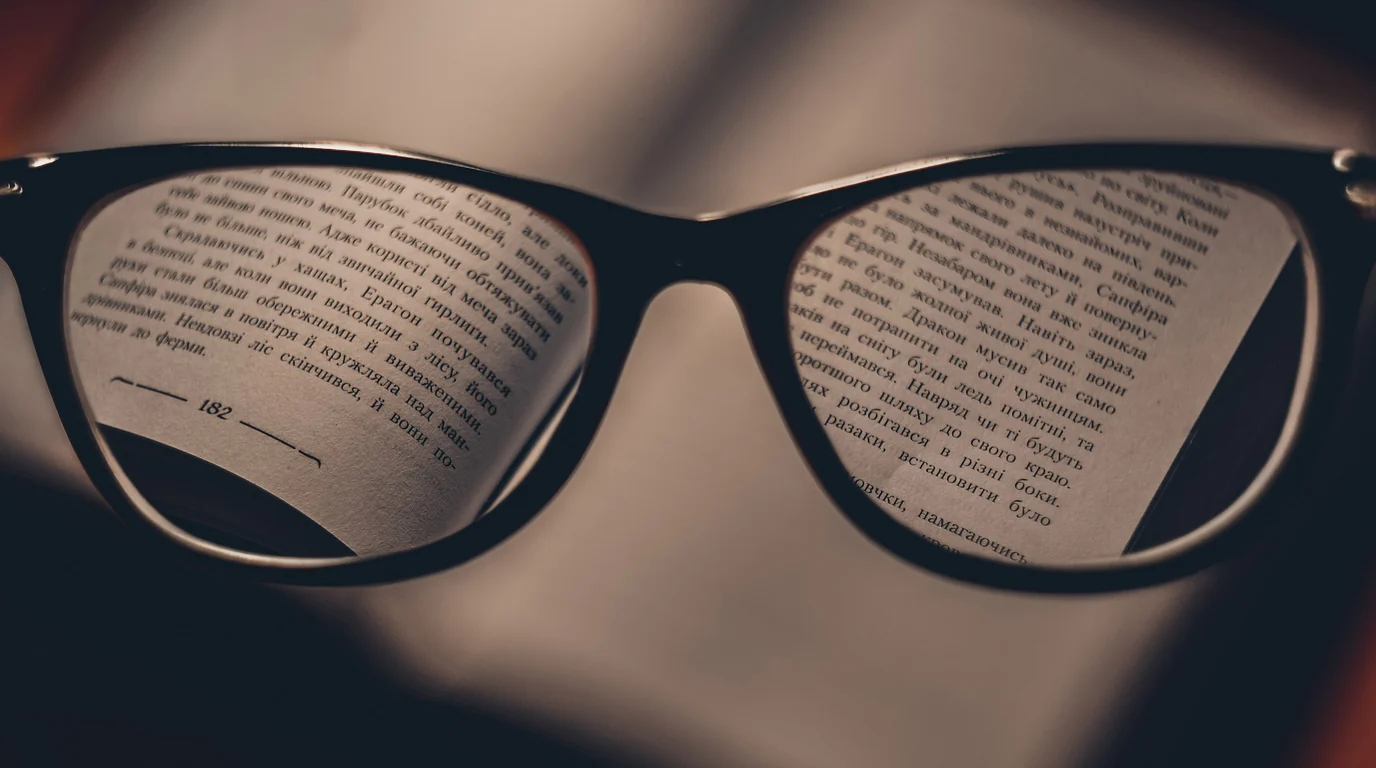 Black reading glasses held above an open book, the printed lines magnified in the lenses.