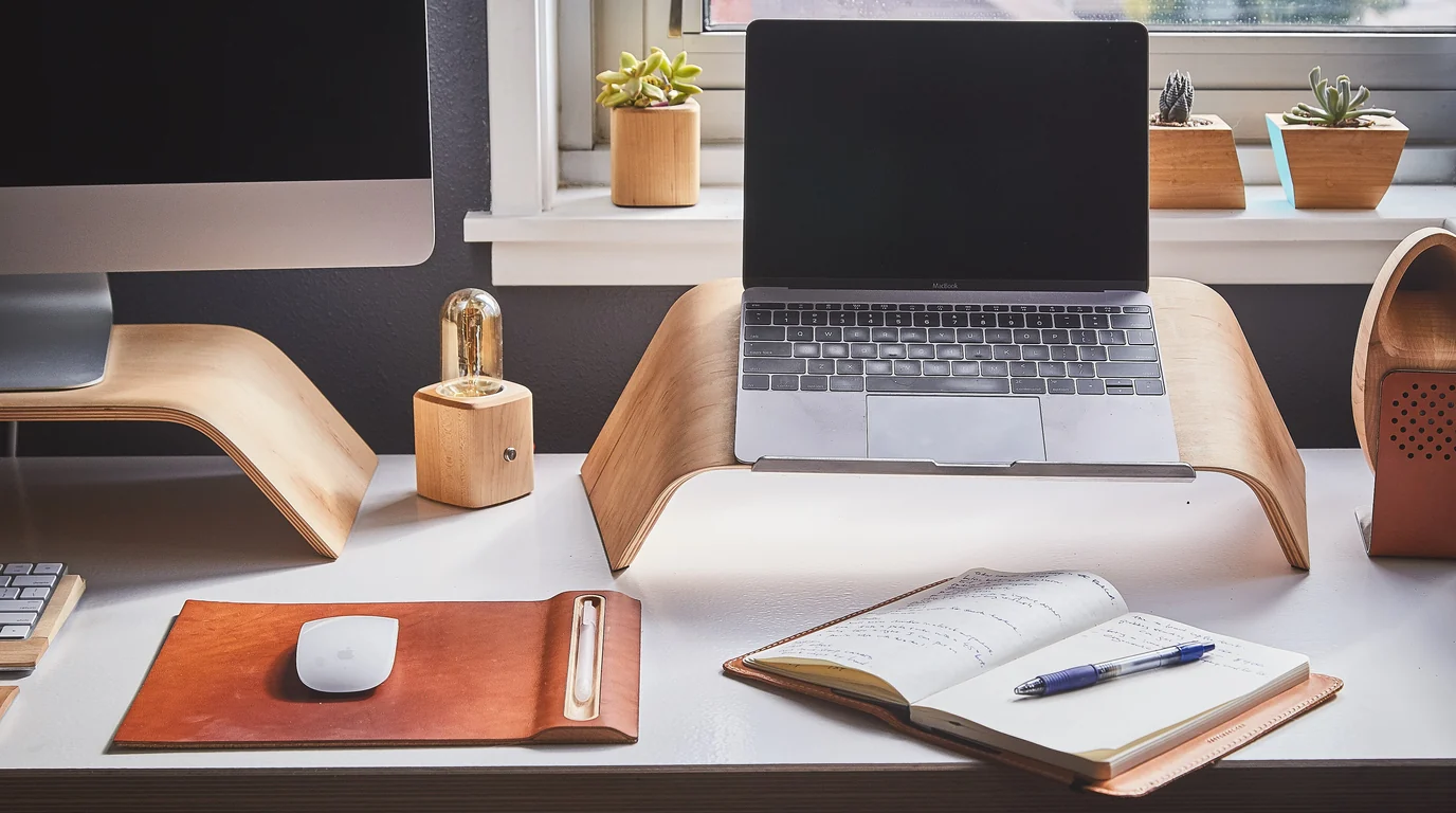A tidy desk with a laptop on a wooden stand, a leather desk mat, a mouse and an open notebook by a window.