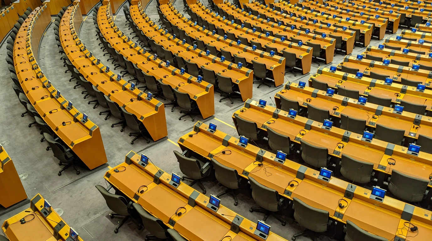 Curved rows of empty yellow desks with small monitors in the European Parliament chamber.