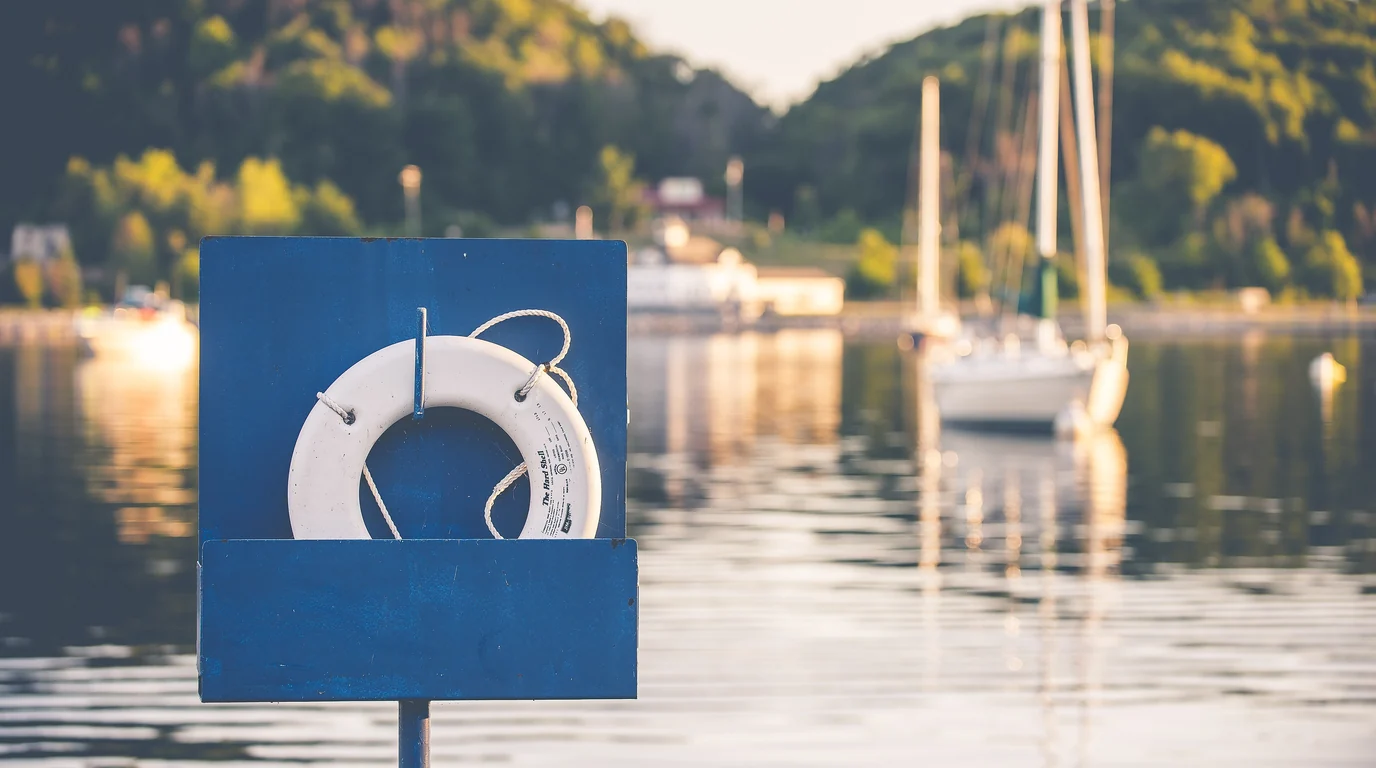 A white lifebuoy in a blue box on a jetty, sailing boats moored on the water behind it.