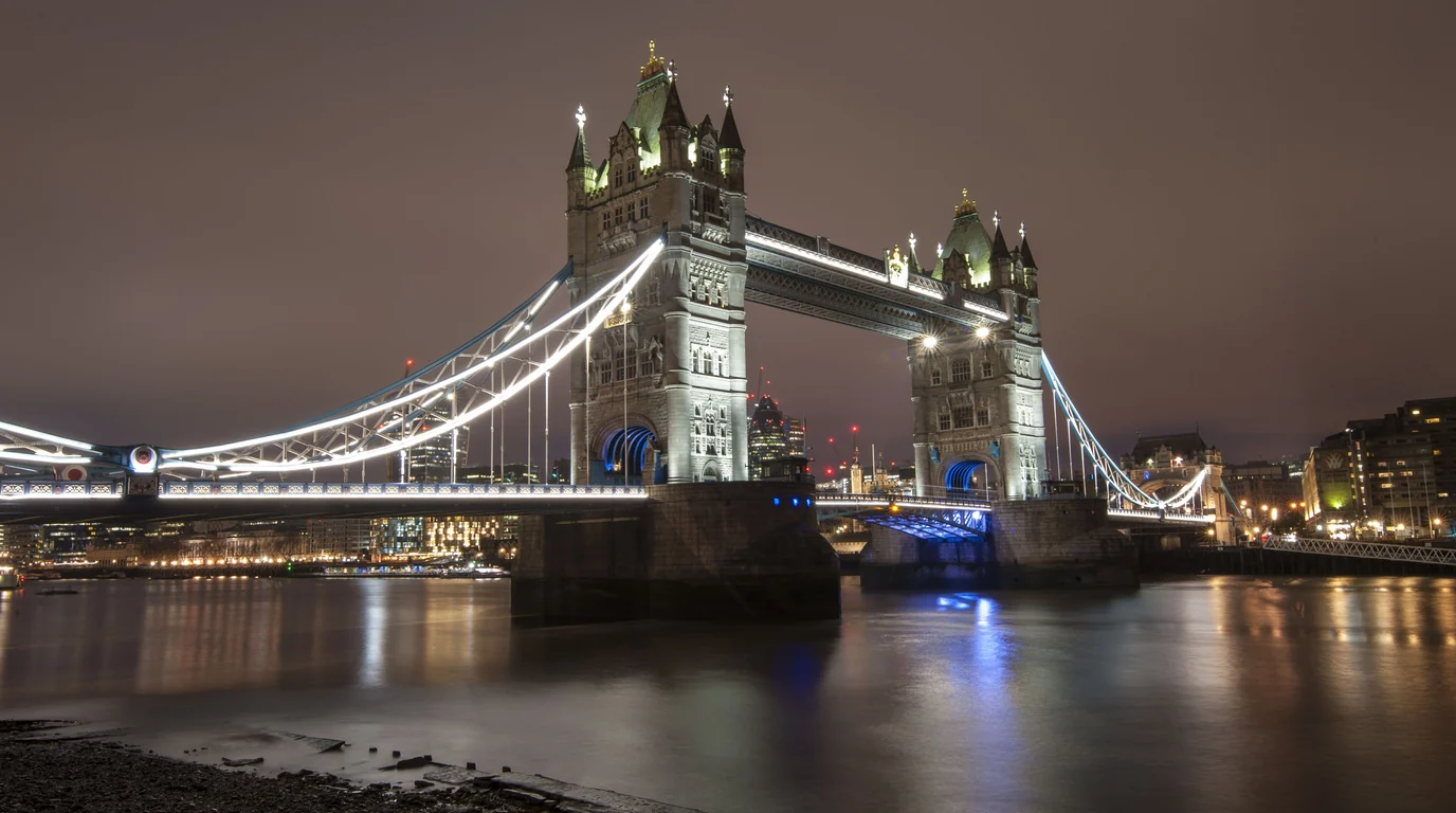 Tower Bridge in London lit up at night, its lights reflected in the Thames.