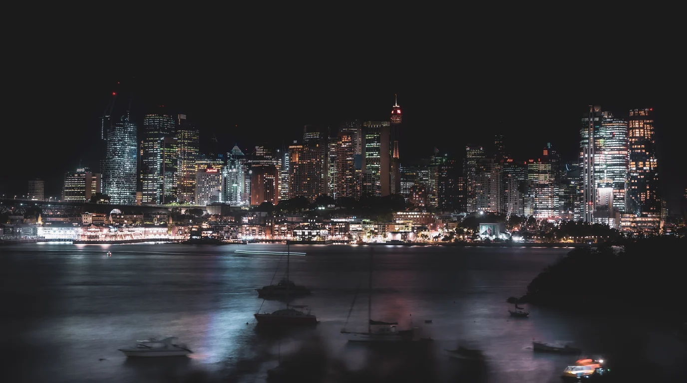 Sydney's lit skyline across the harbour at night, boats moored in the dark water below.