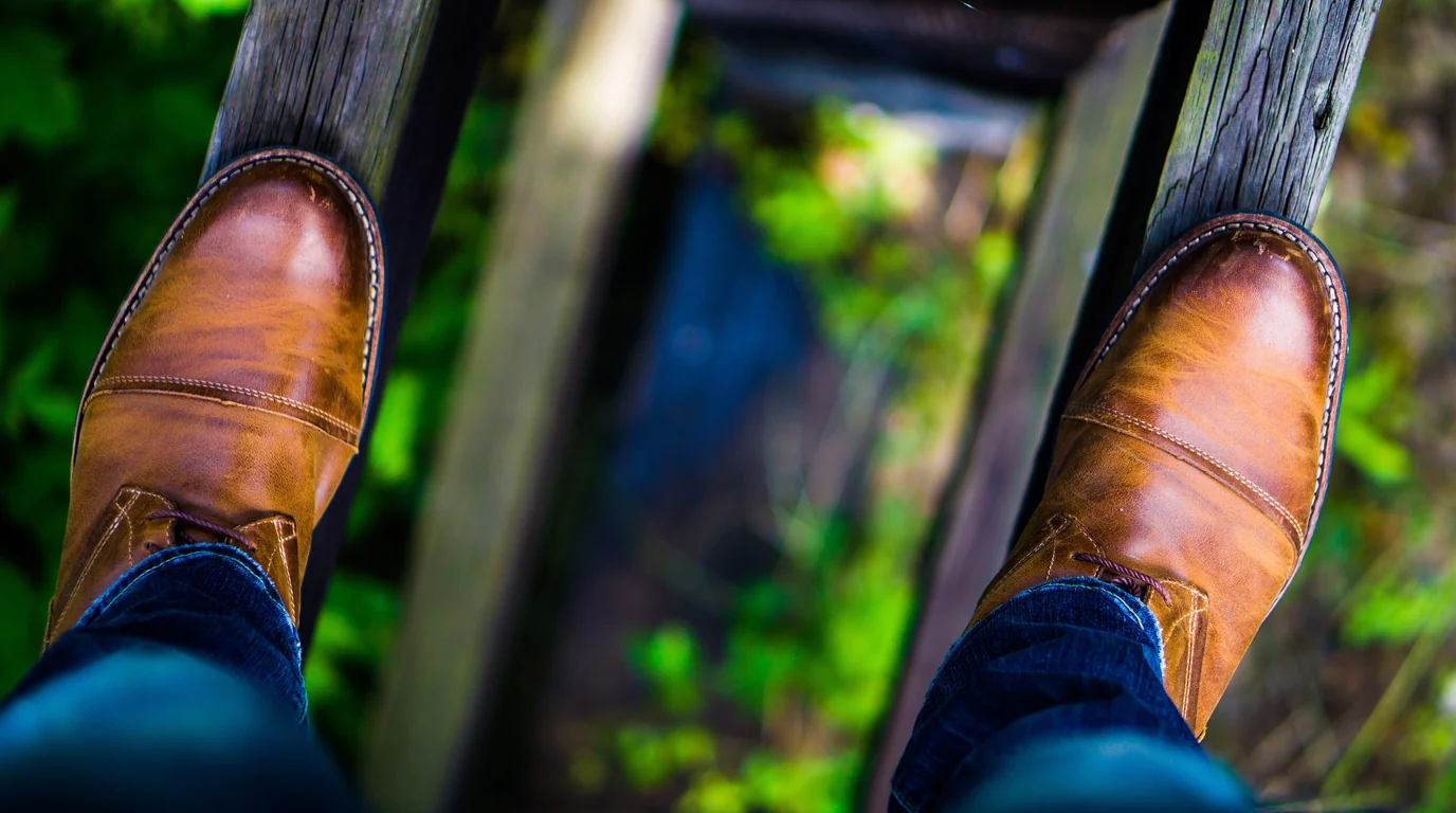 Looking down at a pair of brown leather boots balanced on a narrow wooden beam above greenery.