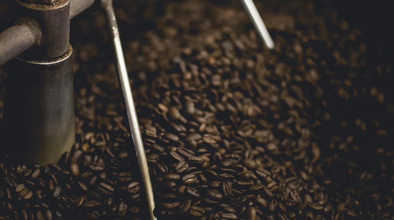 Dark roasted coffee beans turning in a roaster drum, a metal stirring arm cutting through them.